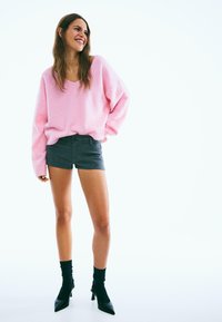 Różowy, oversize'owy sweter, ciemne szorty z denimu, czarne buty na wysokim obcasie i czarne skarpetki do kostki. Tło jednolite białe.