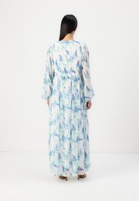 Femme aux longs cheveux foncés portant une robe maxi blanche à imprimé floral bleu avec des manches longues, debout de dos sur un fond blanc uni.
