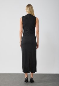 Black sleeveless dress with a fitted design, ruched detailing at the back, and a side slit. Smooth fabric and knee-length cut.