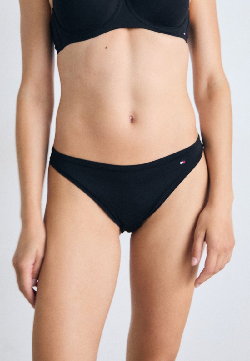 Kvinne iført svart bikini topp og bunn som står mot en ensfarget lys bakgrunn, viser midje og øvre lår.