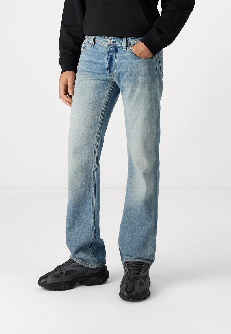 Jeans de perna reta azul claro com um efeito desbotado, cinco bolsos e acessórios em metal. Usados com ténis pretos e um moletom preto.