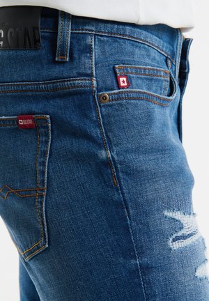 Niebieskie dżinsy charakteryzują się slim fit, z przetartymi akcentami na udach, kontrastowymi szwami oraz naszywkami z logo blisko kieszeni.