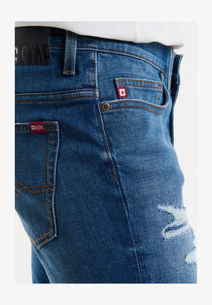 Zilās džinsu bikses ir ar šauru piegriezumu, tām ir nolietoti akcenti uz augšstilbiem, kontrastējoša diega izšuvumi un logo plāksnītes tuvu kabatām.