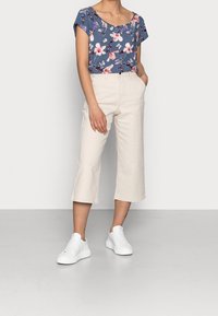 Modrý krátký top s květinovým vzorem v růžové a bílé barvě, doplněný krémovými zkrácenými kalhotami a bílými teniskami.