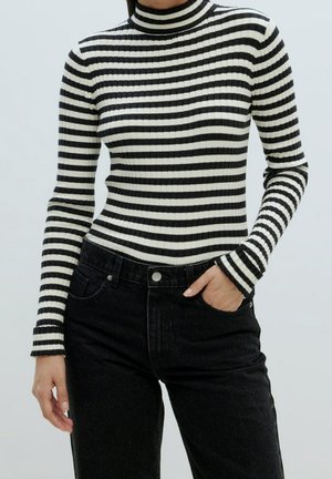 Woman wearing a black and white horizontal striped turtleneck sweater and high-waisted black jeans with one hand in pocket.