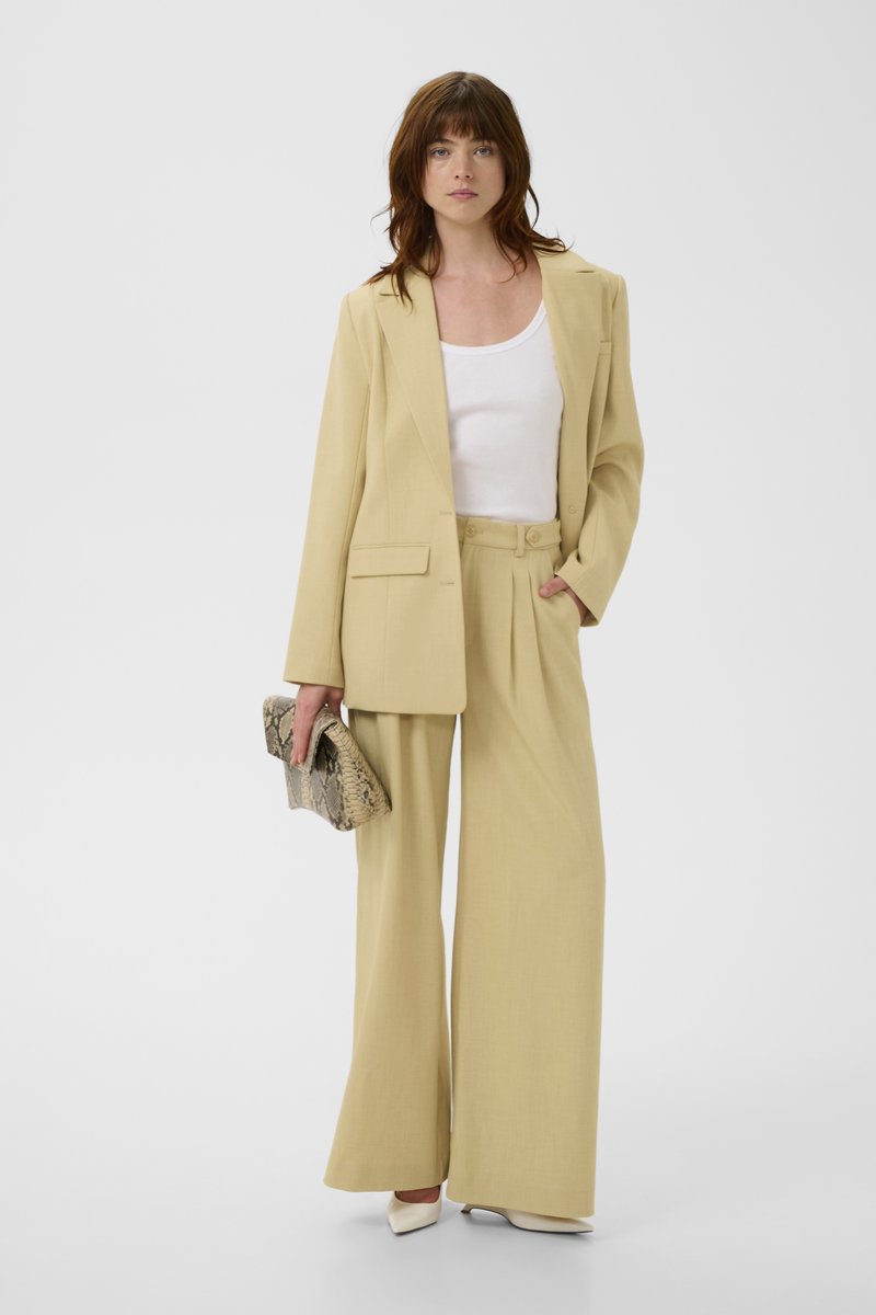 Femme portant un pantalon large beige, un blazer assorti, un haut blanc, des talons blancs, tenant une pochette à motifs sur un fond blanc uni.