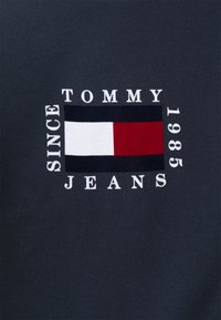 Tummansininen kangas, jossa on valkoinen ja punainen suorakaiteen muotoinen logo, jossa lukee "TOMMY JEANS" ja "SINCE 1985" boldilla valkoisella tekstillä.