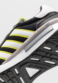 Chaussure de sport noire et blanche avec des accents jaunes ; tige en mesh, col rembourré et semelle en caoutchouc présentant des motifs de traction distincts.