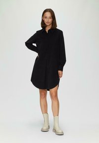 Schwarzes Hemdkleid mit einem kragenförmigen Ausschnitt, langen Ärmeln und abgerundetem Saum. Kombiniert mit hellbeigen Knöchelstiefeln. Glatte Stoffstruktur.