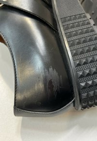 Chaussure en cuir noir avec des marques d'usure visibles sur le côté et une semelle en caoutchouc épaisse et texturée avec des motifs triangulaires.