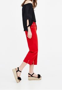 Schwarzes schulterfreies Oberteil, rote Cropped-Jeans, schwarze Plateausandalen mit einem Pompon-Akzent und geflochtenen Jutesohlen. Minimalistisches Design und kontrastierende Farben.