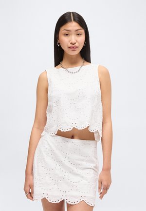 Młoda kobieta z długimi ciemnymi włosami, ubrana w biały, bezrękawnik z ażurowym wzorem i dopasowaną spódnicę, uzupełniona srebrną biżuterią.