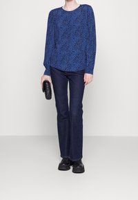 Niebieski top z długim rękawem o teksturowanym wzorze, w połączeniu z ciemnoniebieskimi dżinsami. Modelka trzyma czarną torebkę i ma na sobie czarne buty.