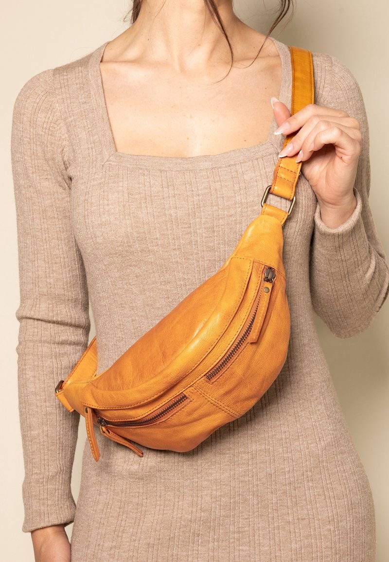 Femme portant une robe-pull beige côtelée tenant un sac en cuir camel porté en bandoulière avec deux compartiments zippés visibles.