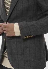 Blazer gris con un patrón, confeccionado en un tejido texturizado, que cuenta con un bolsillo en el pecho y cuatro botones dorados en la manga. Combinado con un suéter color crema.