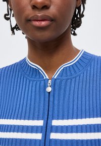 Κοντινό πλάνο ενός ατόμου που φοράει μπλε ριμπ πλεκτή ζακέτα με φερμουάρ, λευκές ρίγες και μικρό στρογγυλό τράβηγμα φερμουάρ.