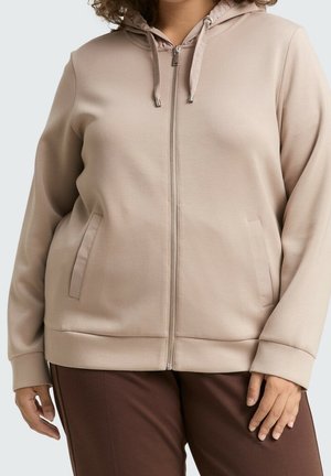 Osoba ubrana w beżową bluzę na zamek z kieszeniami z przodu i sznurkami, w połączeniu z ciemnobrązowymi spodniami, na jednolitym tle.