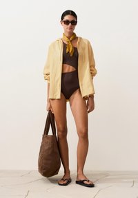 Femme portant un maillot de bain marron foncé à découpes, une veste zippée jaune clair, une écharpe jaune, des lunettes de soleil noires, des sandales noires et portant un grand sac cabas marron.