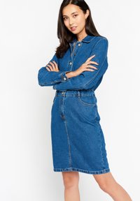 Denim srajčna obleka v srednjem pranju modre barve, ki ima ovratnik, sprednje gumbe, dolge rokave, zategnjeno pasovno območje in krilo do kolen.