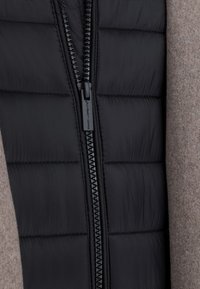 Czarna kamizelka puchowa o pikowanej fakturze i błyszczącym wykończeniu, z centralnym zapięciem na zamek oraz brązowymi akcentami z wełny na bokach.