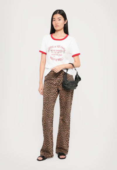 Balta T-kreklu ar sarkaniem akcentiem un grafisko apdruku, apvienota ar brūnu leopardu raksta biksēm. Modelis tur melnu, studētu rokassomu.