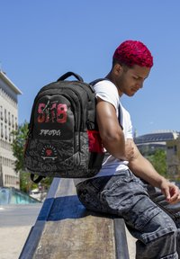 Mochila negra con acentos rojos y gráficos de estilo graffiti. Tiene correas acolchadas, múltiples compartimentos y un bolsillo lateral de malla.