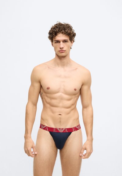 Tanga azul-marinho com uma cintura elástica vermelha apresentando um padrão de logótipo. O material parece suave, com design ajustado realçado.