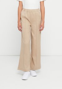 Kvinne iført beige vide bukser med elastisk midjebånd og hvite joggesko, som står mot en enkel lys bakgrunn.