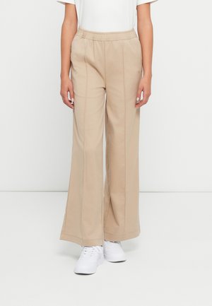 Kvinne iført beige vide bukser med elastisk midjebånd og hvite joggesko, som står mot en enkel lys bakgrunn.