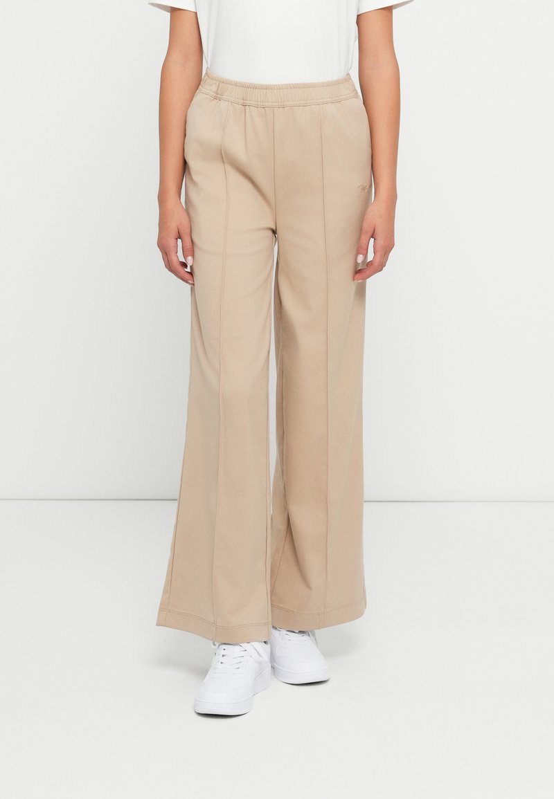 Kvinne iført beige vide bukser med elastisk midjebånd og hvite joggesko, som står mot en enkel lys bakgrunn.