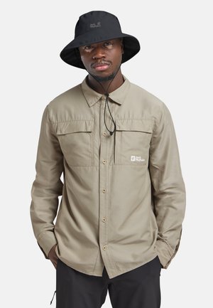Uomo che indossa una camicia beige con bottoni e tasche con patta e un cappello a secchiello nero con cinturino sotto il mento, in piedi con le mani nelle tasche.