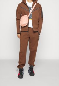 Person som bär en brun Nike-träningsdräkt, vit Nike-t-shirt, svarta sneakers med röda och blå detaljer, samt en rosa Nike-messengerväska.