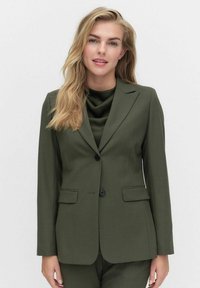 Olivgrüner maßgeschneiderter Blazer aus strukturiertem Stoff mit einem knopfartigen Verschluss, zwei vorderen Taschen und Reverskragen. Kombiniert mit einem fließenden grünen Top.