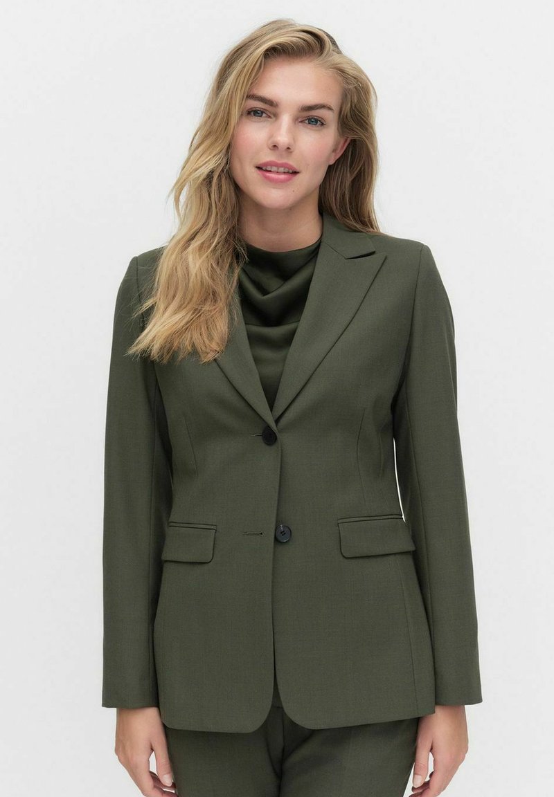 Olivgrüner maßgeschneiderter Blazer aus strukturiertem Stoff mit einem knopfartigen Verschluss, zwei vorderen Taschen und Reverskragen. Kombiniert mit einem fließenden grünen Top.