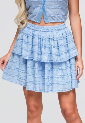 Femme portant une jupe bleu clair à volants et un crop top assorti, montrant le ventre et une partie des jambes, debout, les mains détendues.