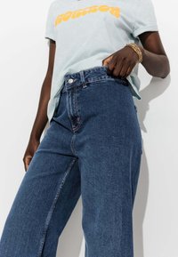 Jean en denim ample de couleur bleu foncé avec des coutures visibles, taille haute et fermeture à bouton en métal ; associé à un t-shirt graphique bleu clair.