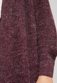 Gros plan sur une personne portant un pull en maille texturée de couleur violet foncé avec un poignet côtelé visible au niveau du poignet.