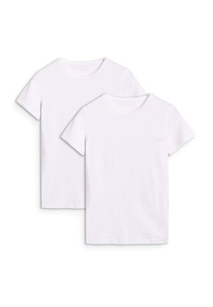 Młoda kobieta w czapce z daszkiem, białym t-shircie, jasnoszarych spodniach dresowych i szarych trampkach, stojąca z jedną ręką w kieszeni na jednolitym jasnym tle.