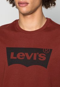 Mørkerød bomulds t-shirt med et stort sort Levi's logo i en buet stil på tværs af brystet. Rund halsudskæring og korte ærmer.