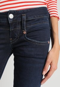 Close-up of person wearing dark blue jeans and a red and white striped long-sleeve shirt, hand resting by the thigh.