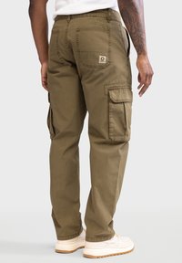 Olivgrüne Cargo-Hosen mit lockerer Passform, ausgestattet mit seitlichen Taschen, Gesäßtaschen und einem gewebten Logo-Patch auf der rechten Tasche. Sichtbare Nähte setzen Akzente.