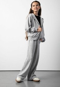 Szara bluza z kapturem na zamek błyskawiczny z elastycznymi mankietami oraz dopasowane luźne spodnie dresowe. Obydwa ubrania wykonane są z miękkiego materiału, noszone z białymi sneakersami z brązowymi podeszwami.