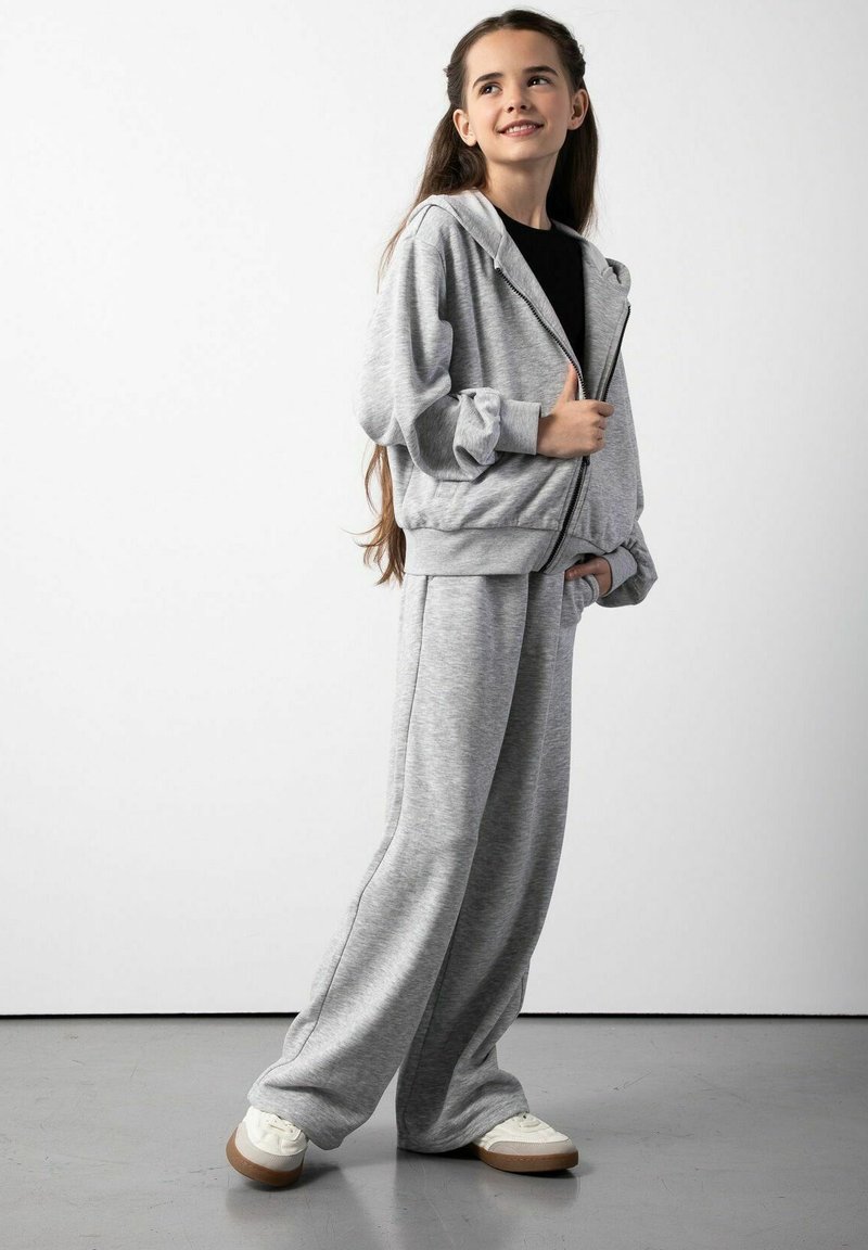 Szara bluza z kapturem na zamek błyskawiczny z elastycznymi mankietami oraz dopasowane luźne spodnie dresowe. Obydwa ubrania wykonane są z miękkiego materiału, noszone z białymi sneakersami z brązowymi podeszwami.