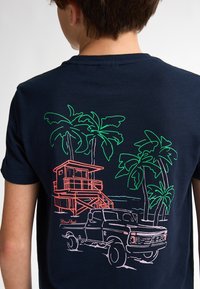 Námornická modrá tričko s grafickým designem, který zobrazuje růžovou plážovou budku, zelené palmy a bílý vintage vůz, vše obkreslené neonovými barvami.