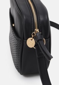 Zwarte leren crossbodytas met een geweven textuur, gouden ritssluiting en charm-detail. De tas heeft een gestructureerde vorm en een platte riem.