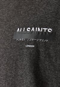 T-shirt en textile gris avec le texte brodé en blanc "ALLSAINTS" et une bande texturée noire masquant du texte supplémentaire. Design simple à col rond.