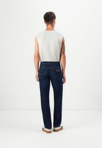 Jeans rectos de color azul oscuro, de material de algodón, con bolsillos traseros que presentan un patrón de costura distintivo y una etiqueta de cuero marrón.