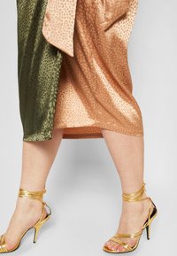 Robe en satin avec un imprimé feuillage dans des tons verts et pêches, associée à des sandales à talons hauts dorées à lanières, présentant un design de chevillère.