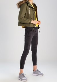 Olivově zelená zkrácená bunda s kožešinovou podšívkou na kapuci, žlutý top pod tím, černé vypasované kalhoty se zipovými detaily a šedé sportovní boty.