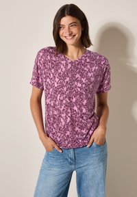 T-shirt à imprimé graphique violet et rose avec des manches courtes et un col en V. Tissu doux, coupe décontractée, associé à un jean bleu clair.